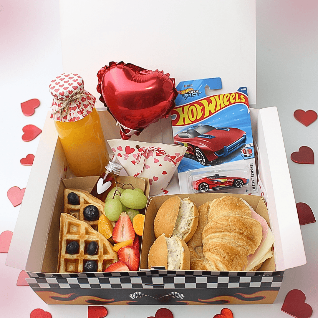Desayuno Amor sobre ruedas. Caja de desayuno temática Hot Wheels abierta con wafles, frutas, croissant, sándwiches, jugo, un carrito Hot Wheels azul y rojo, y un globo de corazón metálico sobre fondo blanco con corazones