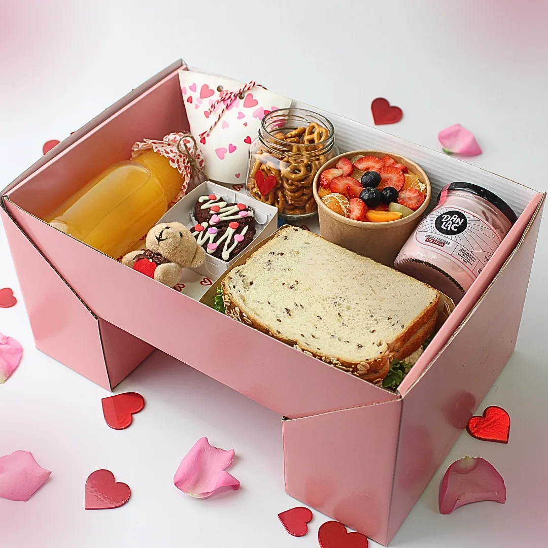 Desayuno Picnic de Amor Premium es una caja de desayuno romántica abierta de color rosa con tapa convertible estilo picnic, llena de productos para aniversario. Incluye sándwich multigrano, ensalada de frutas, jugo, pretzels de corazón en frasco, yogurt Danlac, chocolates, oso de peluche y tarjeta personalizada